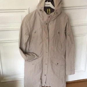 Beige vår kappa från Lindex - Säljer en stilren beige kappa från Lindex med huva och dragkedja. Jackan har långa ärmar och flera praktiska fickor med knappar. Perfekt för kyligare dagar.