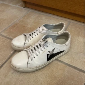 Vita sneakers från Axel Arigato - Snygga vita sneakers från Axel Arigato med en svart fågel på sidan. Stilren sko som passar perfekt till vardags. Jag tror att jag har kvar box och påse. Inte defekter som påverkar skon