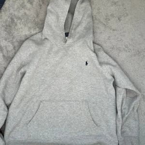 Grå hoodie från Ralph Lauren - Säljer en stilren grå hoodie från Ralph Lauren med klassisk känguruficka och broderad logga på bröstet. Perfekt för en avslappnad look. Huvan har ett mjukt foder för extra komfort.