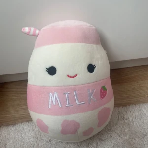 Squishmallows mjukisdjur mjölkflaska - Säljer en söt Squishmallows mjukisdjur formad som en mjölkflaska. Den har en rosa och vit design med ordet 'MILK' broderat och en liten jordgubbe. Perfekt för kramar och dekoration! 🍼🍓