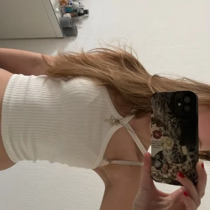 Vit topp från Gina Tricot - Säljer en stilren vit ribbad linnetopp från Gina Tricot. Toppen har en dekorativ stjärndetalj framtill och smala axelband. Perfekt för varma dagar eller som ett lager under en jacka.