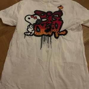 Vit t-shirt med Snoopy-tryck från H&M - Säljer en vit t-shirt från H&M med ett coolt Snoopy-tryck på både fram- och baksidan. T-shirten har en normal passform och är kortärmad, perfekt för en avslappnad stil. Trycket på baksidan har en graffiti-inspirerad design med texten 'Big Deal'.