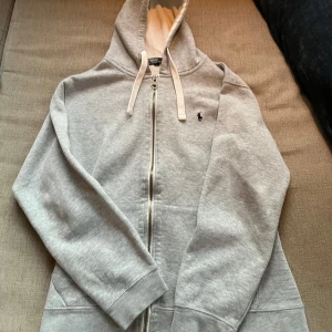 Grå hoodie från Polo Ralph Lauren - Säljer en stilren grå hoodie från Polo Ralph Lauren med dragkedja och klassisk logga på bröstet. Tröjan har en mjuk insida och justerbar huva med vita snören. Perfekt för en Laidback look.