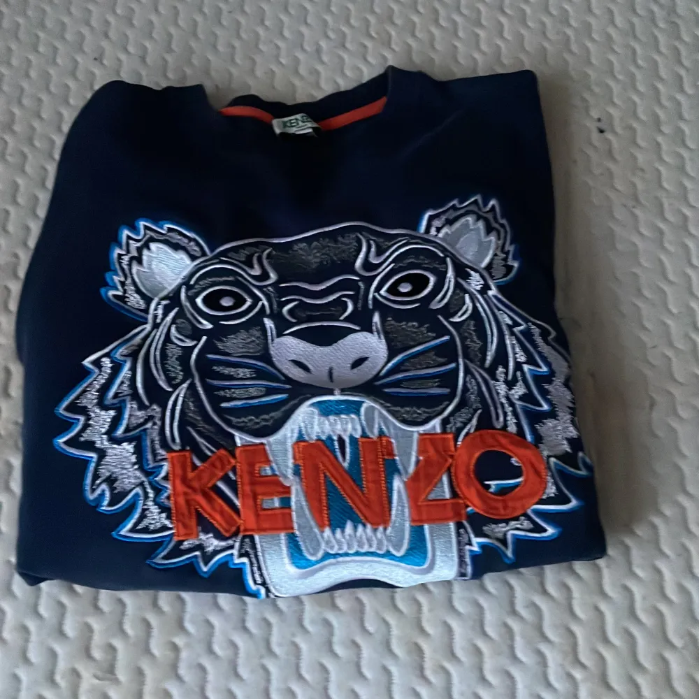 Säljer en mörkblå sweatshirt från Kenzo med ett stort tigerbroderi och logotyp på framsidan. Tröjan har en normal passform och är tillverkad i ett mjukt material. Perfekt för en stilren och avslappnad look.tröjan är knappt använd har haft på mig den kanske fem gånger.det är small i barn storlek.. Neuletakit & Villapaidat.