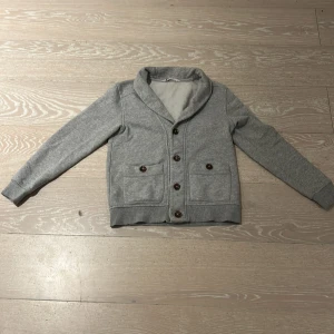 Grå kofta med knappar - Säljer en stilren grå kofta med knappar framtill. Den har en mjuk krage och långa ärmar, perfekt för kyligare dagar. Koftan har två fickor med knappar som ger en extra touch. Passar bra till både vardag och mer uppklädda tillfällen.