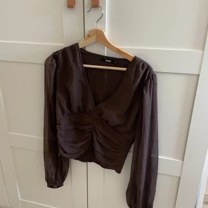 Brun blus från Bik Bok - Säljer en elegant brun blus från Bik Bok med v-ringning och rynkade detaljer framtill. Blusen har långa ärmar och en stilren design som passar perfekt för höstgarderoben.