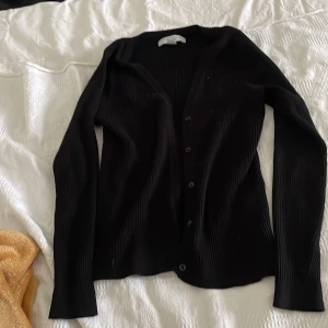 Svart ribbad kofta från Amisu - Säljer en stilren svart ribbad kofta från Amisu. Koftan har långa ärmar och knappar framtill, perfekt för lager-på-lager-stil. Passar både till vardags och mer uppklädda tillfällen.