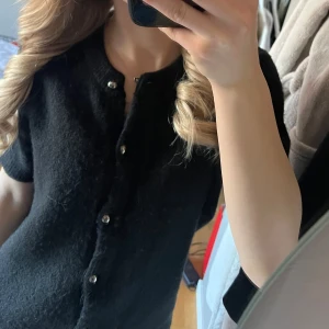 Svart kortärmad kofta med knappar från Gina  - Säljer en stilren svart kofta med korta ärmar och knappar framtill. Perfekt för lager-på-lager och passar till många olika outfits. Från Gina, nypris 399kr