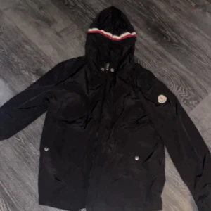 Svart vindjacka från Moncler - Säljer en stilren svart vindjacka från Moncler med huva och dragkedja. Jackan har långa ärmar och en snygg randig detalj i rött och vitt och blå på huvan. Perfekt för blåsiga dagar!