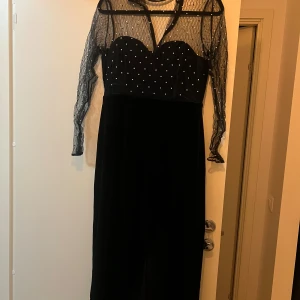 Svart klänning med mesh och pärlor - Elegant svart klänning med långärmad mesh-överdel och pärldetaljer. Klänningen har en åtsittande passform upptill med ett hjärtformat liv och transparenta ärmar. Perfekt för en festlig kväll!