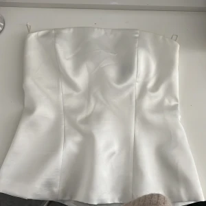 Vit satin korsettopp från Zara - Supersnygg vit korsettopp från Zara i glansigt satinliknande material. Toppen är axelbandslös och har en figurnära passform med markerade sömmar och dragkedja bak. Perfekt för att skapa en clean och chic look. Använd endast en gång för att den var för stor för mig.