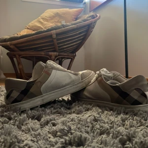 Burberry rutig sneakers i skinn - INTRESSE KOLL! Snygga vita sneakers från Burberry med klassiskt rutigt mönster i beige, svart och brunt på hälen. Skorna har rund tå, snörning och platt sula. Tillverkade i skinn med detaljer i syntet. Perfekta för dig som gillar lyxiga street vibes.