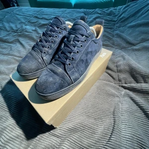 Blå mocka sneakers från Louboutin - Snygga sneakers från Christian Louboutin i mörkblå mocka med klassisk röd sula. Skorna har rund tå, snörning och diskreta detaljer. Insidan är ljusbeige och de levereras med originalkartong och dustbag. Perfekta för dig som vill sticka ut med exklusiva sneakers.