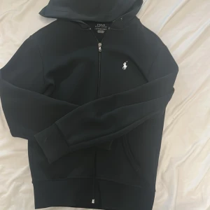 Svart hoodie från Polo Ralph Lauren - Svart hoodie från Polo Ralph Lauren med klassisk vit broderad logga på bröstet. Tröjan har dragkedja framtill, huva och två fickor. Tillverkad i mjuk bomullsblandning och har långa ärmar med ribbade muddar. Perfekt för en chill och stilren look.