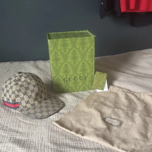 Beige gucci keps - Säljer en snygg beige keps från Gucci med klassiskt GG-monogram i mörkblått. Kepsen har en böjd skärm, justerbar rem bak och en röd-blå rand på sidan. Materialet är bomull och polyester, insidan är mörkblå. Kommer med originalkartong och dustbag.