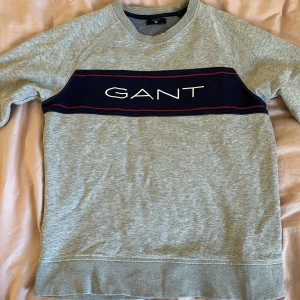 Grå sweatshirt från GANT med logga - Skick 8/10. storlek S. Snygg grå sweatshirt från GANT med klassisk marinblå panel och vit broderad logga över bröstet. Tröjan har rund hals och långa ärmar. 