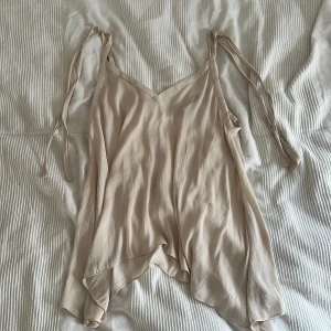 Beige linnetopp med knytband - Säljer en luftig beige linnetopp med smala axelband som knyts på axlarna. Toppen har v-ringning och ett flowy, asymmetriskt fall. Perfekt för varma dagar och enkel att styla till olika looks.