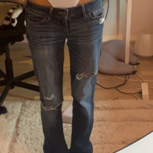 Blå bootcut jeans från Hollister - Säljer ett par snygga blå jeans från Hollister med bootcut passform och slitna detaljer på benen. Jeansen har klassisk femficksdesign, låg midja och coola slitningar både fram och bak. Materialet är mjukt denim och de har orangea sömmar på bakfickorna.