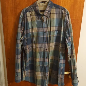 Rutig blå skjorta från Dressmann - Snygg rutig skjorta från Dressmann i blå och beige toner. Skjortan har klassisk krage, knäppning framtill och en bröstficka. Tillverkad i mjuk bomull och har långa ärmar med manschettknappar. Perfekt för en avslappnad men stilren look.