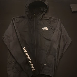 Svart vindjacka från The North Face - Svart vindjacka från The North Face med huva och hel dragkedja. Jackan har logga på bröstet och ärmen, samt elastiska muddar. Tillverkad i lätt polyester, perfekt för blåsiga dagar. Snygg och enkel design med sportig vibe.