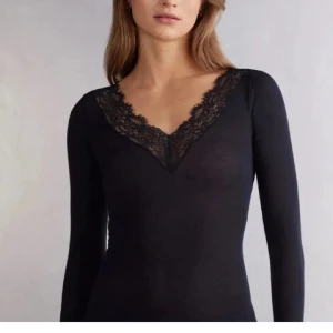 Intimissimi långärmad  - Supersnygg svart långärmad topp med v-ringning och spetsdetaljer runt halsen. Tight passform som framhäver figuren och ger en chic vibe. Perfekt att matcha med jeans eller kjol för en trendig look. Materialet känns mjukt och följsamt mot huden.