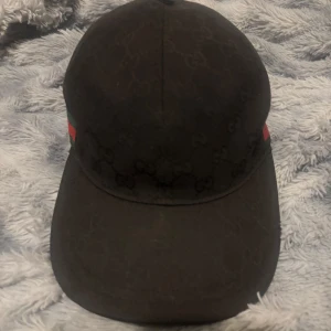 Gucci keps (ÄKTA) Svart - Snygg svart keps från Gucci med klassiskt GG-monogram i tyget och ikoniska gröna och röda ränder på sidorna. Justerbar rem baktill och böjd skärm. Perfekt accessoar för dig som vill sticka ut med lyxig streetstyle.  Storlek M och lappen är klippt för det störde mig men den är äkta 