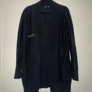 Svart lång kofta med fickor - Mysig svart lång kofta med bred ribbad kant och stora fickor framtill. Perfekt att slänga på sig över en t-shirt eller topp. Koftan har lång ärm och en loose passform som ger en avslappnad vibe. Inga fläckar, kameran fick med ett vitt ljus.