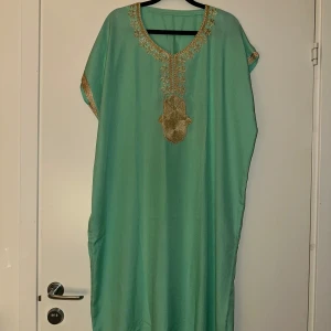 Mintgrön långklänning med gulddekor - Snygg mintgrön långklänning med v-ringning och vackra guldfärgade broderier runt halsen och på ärmarna. Löst sittande modell med korta ärmar, perfekt för dig som gillar en avslappnad och elegant stil.