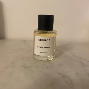 ESSNCE Dusty Amber Eau de Parfum - En stilren parfymflaska med svart lock och transparent glas, fylld med en ljusgul doftvätska. Dusty Amber är en Eau de Parfum med minimalistisk etikett och modern känsla, perfekt för dig som gillar eleganta och diskreta dofter.