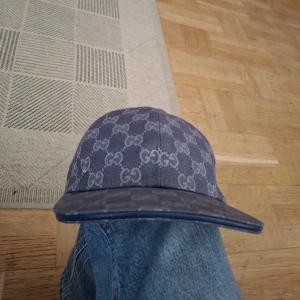 Blå Gucci keps med GG-mönster - Snygg blå keps från Gucci med klassiskt GG-monogram över hela kepsen. Justerbar rem baktill i blått läder och böjd skärm. Tillverkad i bomullsmaterial med exklusiv känsla. Perfekt för dig som vill sticka ut med en lyxig accessoar.