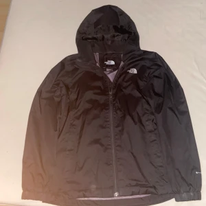 Svart vindjacka från The North Face - Svart vindjacka från The North Face med huva och hel dragkedja framtill. Jackan har meshfoder på insidan och logga på bröstet. Perfekt för blåsiga dagar och enkel att slänga på sig när vädret svänger.