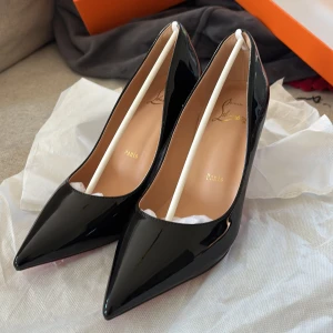 Svarta lackpumps från Louboutin - Klassiska svarta pumps i lack från Christian Louboutin med ikonisk röd sula. Skorna har spetsig tå och hög klack, vilket ger en elegant och tidlös look. Perfekta för dig som vill sticka ut med exklusiv design och lyxig känsla.