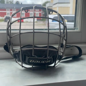 Bauer hockeygaller - Säljer ett Bauer hockeygaller i silverfärgad metall med svart hakskydd och justerbara remmar. Gallret har klassisk burform och passar perfekt för dig som spelar hockey och vill ha bra skydd för ansiktet. Snyggt och funktionellt till hjälmen. storlek på gallret är M.