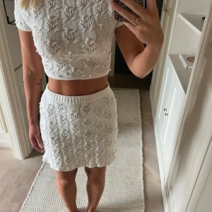 Vit stickad set med pärlor - Supersöt vit stickad kortkjol och top med dekorativa pärlor och ett snyggt hålmönster. Kjolen har en elastisk midja och är gjord i mjukt stickat material och toppen knyter men i ryggen. Perfekt för dig som gillar unika detaljer och vill sticka ut lite extra.