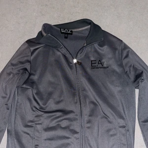 Grå kofta från EA7 Emporio Armani - Snygg Mörkgrå kofta från EA7 Emporio Armani med stilren design och logga på bröstet. Koftan  har dragkedja framtill, ståkrage och två fickor. Perfekt för dig som gillar sportig och clean look. Materialet är lätt och känns skönt mot kroppen.