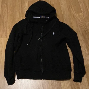 Svart hoodie från Polo Ralph Lauren - Svart hoodie från Polo Ralph Lauren i custom fit. Klassisk design med dragkedja framtill, huva med snörning och vit broderad logga på bröstet. Tillverkad i mjuk bomull, perfekt för en chill och stilren look.