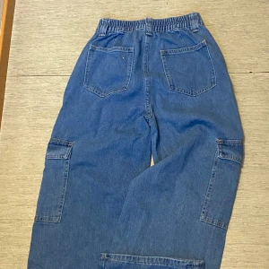 Blå denim cargopants med resårmidja - Säljer ett par blå cargopants i jeansmaterial med breda ben och resår i midjan. Byxorna har stora fickor på sidorna med lock och knapp, samt två bakfickor. Perfekta för en avslappnad och trendig streetstil.