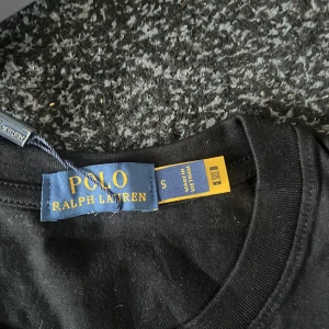 Svart t-shirt Polo Ralph Lauren S - Svart t-shirt från Polo Ralph Lauren i storlek S finns M också . Klassisk rund hals och korta ärmar. På bröstet finns den ikoniska röda Polo-loggan broderad. T-shirten är gjord i mjuk bomull och har en clean, stilren look som funkar till allt. Priset kan diskuteras 
