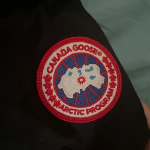 Svart dunjacka från Canada Goose S/P - Svart dunjacka från Canada Goose med klassisk Arctic Program-märke på ärmen. Jackan har en stilren design, dragkedja framtill och är tillverkad i slitstarkt syntetmaterial med dunfyllning. Perfekt för kalla dagar och har en snygg, tidlös look.  Finns lite små fel på den man enkelt kan fixa hemma bara man har tråd och nål helt perfekt och skön annars såklart äkta 