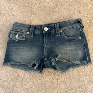 True Religion Joey Cut-Off jeansshorts - Snygga blå jeansshorts från True Religion, modell Joey Cut-Off med låg midja. Klassisk femficksdesign, fransiga kanter och coola detaljer på bakfickorna. Silverfärgade knappar och etikett bak. Aldrig använda! Pris kan diskuteras💕