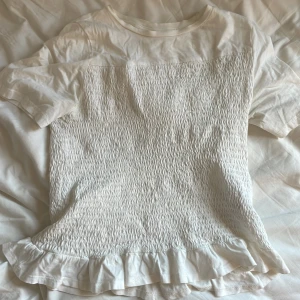 Vit volangtopp från Gina Tricot - Supersöt vit topp från Gina Tricot med smockad framsida och baksida. Toppen har korta ärmar, rund halsringning och en volangkant längst ner som ger en gullig touch. Perfekt för dig som gillar en feminin och trendig stil.