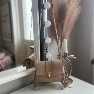 Beige axelväska med fransar och guldkedja - Snygg beige axelväska med guldfärgad kedja och dekorativa fransar på sidorna. Väskan har ett lock med gulddetaljer och vridlås, samt flera fack inuti för smart organisering. Materialet är mockaliknande och ger en lyxig känsla. Super lätt att ta av banden och få en trändig väska lik en kuvert väska