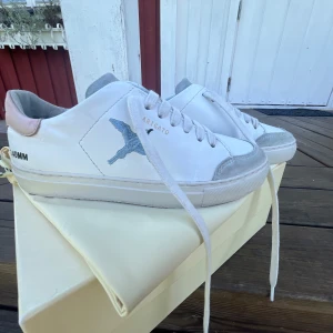 Vit sneaker Axel Arigato med blå fågel - Snygga vita sneakers från Axel Arigato med broderad blå fågel på sidan och ljusgrå mockadetaljer vid tån och plösen. Skorna har ljusrosa hälparti, vita snören och platt sula. Jag har ägt skorna i ett halv år, sulan e lite förstörd men de går ju att köpa en ny. Skriv till mig om ett pris förslag och om fler bilder💓 original priset e 2750kr och som jag kan se finns inte denna modell kvar men SKRIV OM PRIS FÖRSLAG!