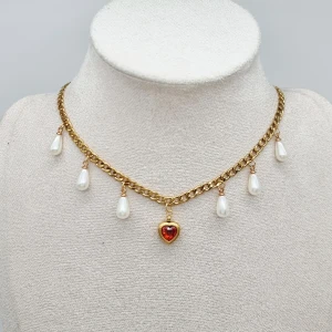 Guldhalsband med pärldroppar & hjärta - Unikt halsband i guldfärgad kedja med sex droppformade vita pärlor och ett rött hjärthänge i mitten. Kedjan har en chunky look och pärlorna hänger snyggt längs halsen. Perfekt statement-piece för dig som gillar romantiska detaljer.