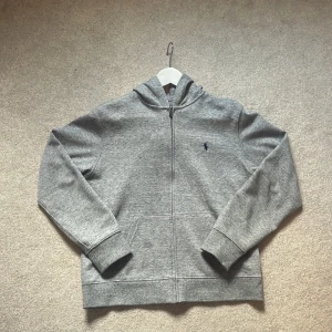 Grå hoodie från Polo Ralph Lauren - Säljer en stilren grå hoodie från Polo Ralph Lauren med dragkedja och huva. Tröjan har broderad logga på bröstet, känguruficka och är tillverkad i mjuk bomullsmix. Perfekt för chill dagar eller när du vill ha en enkel men snygg look.