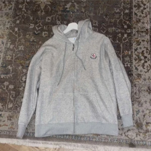 Grå hoodie med dragkedja från Moncler - Snygg grå hoodie från Moncler med dragkedja framtill och huva med snörning. Klassisk logga på bröstet och ribbade muddar vid ärmslut och nederkant. Perfekt för en chill och stilren look.