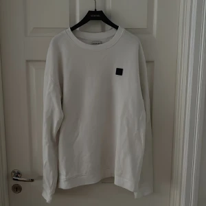 Vit sweatshirt från Acne Studios - Vit sweatshirt från Studio total med klassisk rund hals och svart logopatch på bröstet. Tröjan har en clean look, långa ärmar. Perfekt för dig som gillar minimalistisk stil.