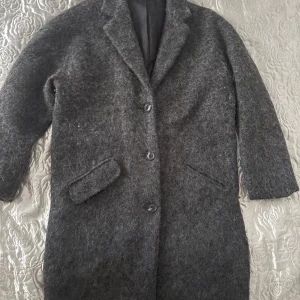 Mörkgrå ullkappa från Gina Tricot - Snygg mörkgrå kappa från Gina Tricot i ullmix med klassisk krage och tre knappar framtill. Kappan har två fickor med lock och en rak, stilren passform som funkar till alla outfits. Perfekt för kyliga dagar när du vill vara både varm och trendig.