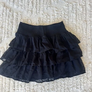 Svart volangkjol från Lindex - Supersöt svart kortkjol från Lindex med flera lager volanger och bred smock i midjan. Kjolen har ett diskret prickigt mönster i tyget och är gjord i ett lätt och luftigt material som ger en härlig känsla. Perfekt för dig som gillar att sticka ut med volanger!