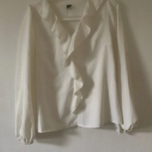 Vit blus med volang och v-ringning - Säljer en stilren vit blus med långärmade, lätt transparenta ärmar och snygg volang längs v-ringningen. Blusen har en luftig passform och är perfekt för dig som gillar en romantisk och trendig look.